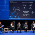 A conference panel on stage during the FF4EuroHPC session. Five speakers sit under a large presentation slide showing project statistics such as partners, experiments, and funding.