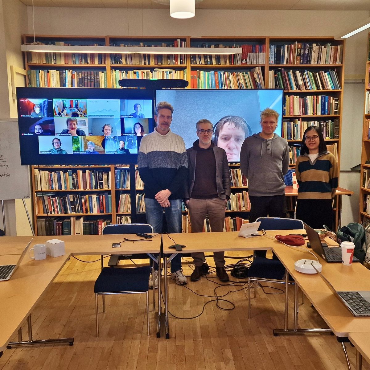 Four people standing in front of two screens, displaying a videocall.