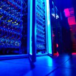 Close-up view of illuminated server racks and cooling tubes inside a high-performance computing data centre, with blue and red lighting reflecting across the floor.