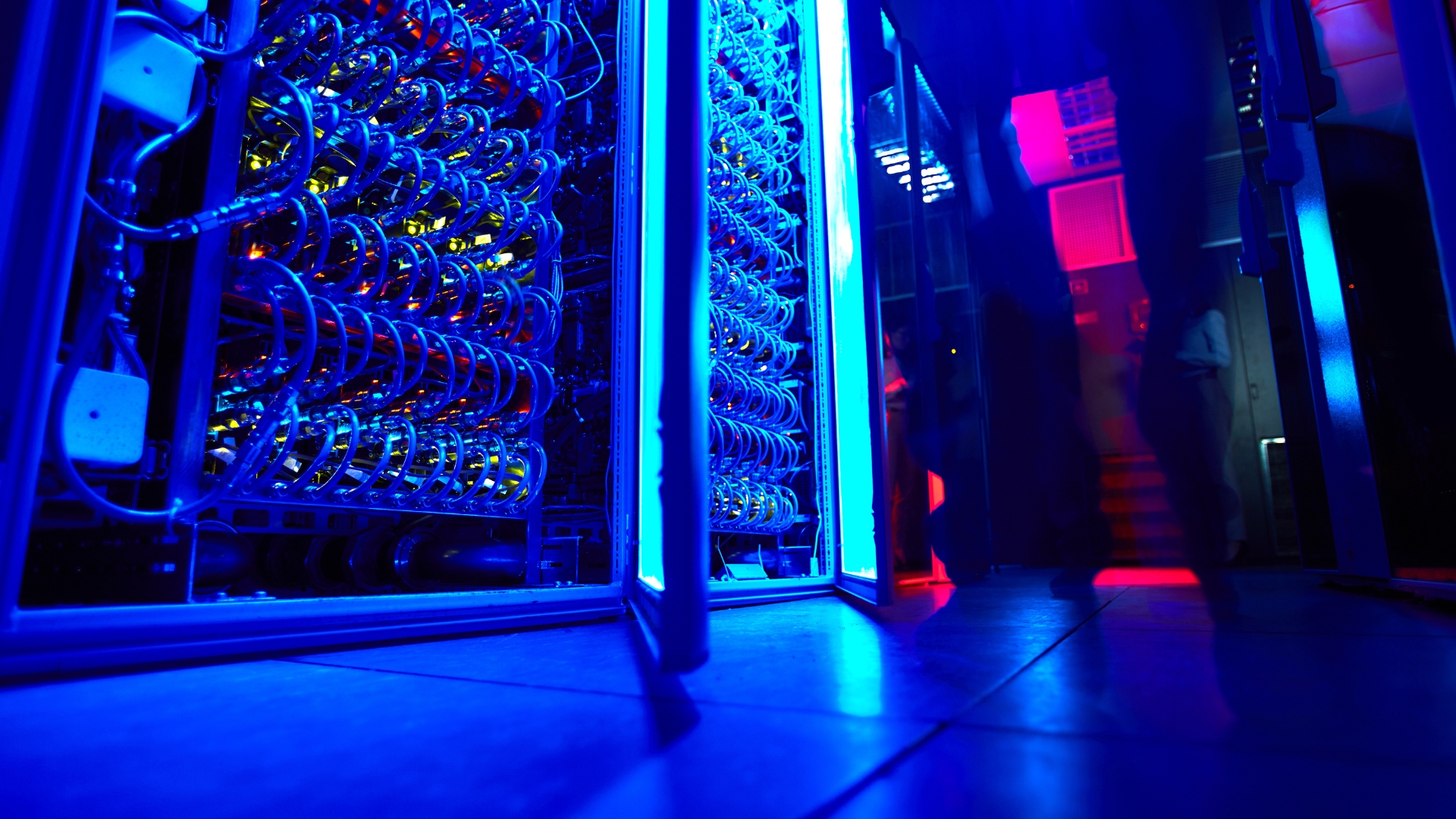 Close-up view of illuminated server racks and cooling tubes inside a high-performance computing data centre, with blue and red lighting reflecting across the floor.