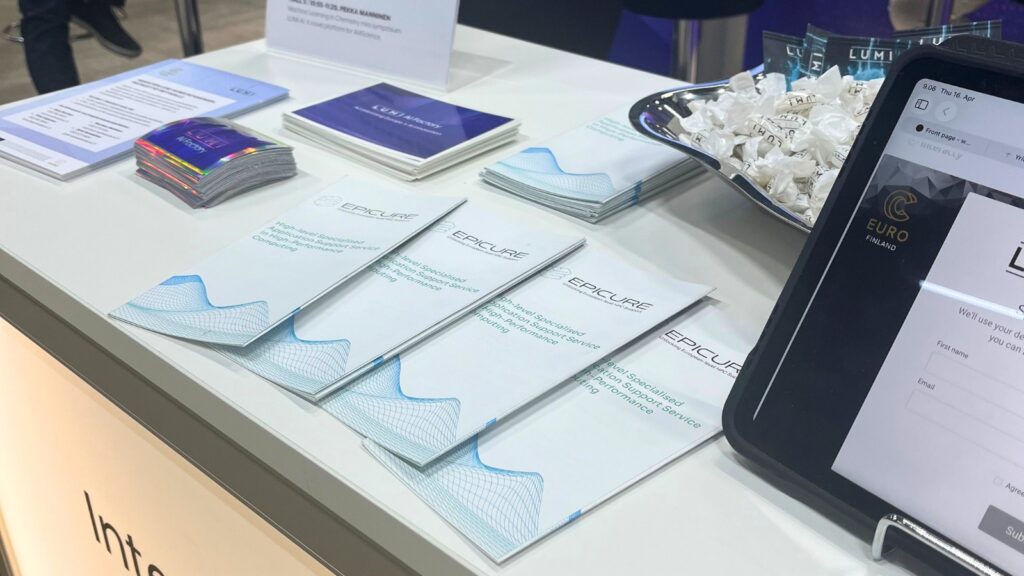 Event stand display with EPICURE brochures, LUMI AI Factory materials, a bowl of wrapped sweets, and a tablet showing a registration form, arranged on a white counter.