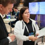 Two attendees at an exhibition stand reviewing a brochure, with EURO Finland and LUMI-related materials visible in the background and a presentation screen behind them.