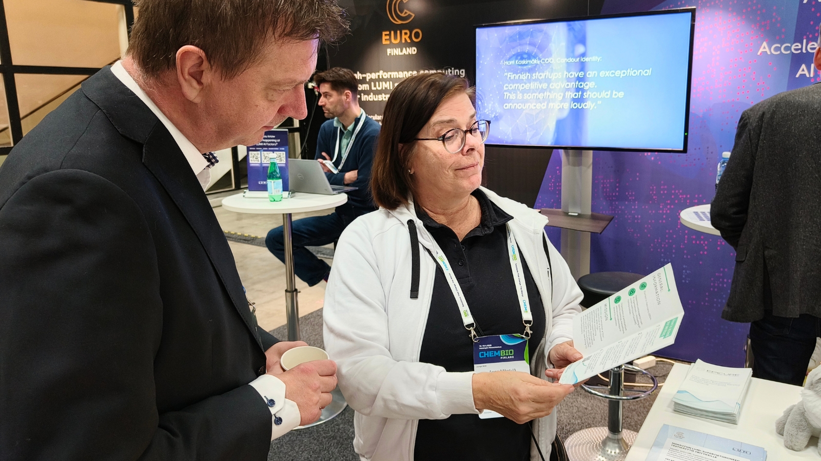 Two attendees at an exhibition stand reviewing a brochure, with EURO Finland and LUMI-related materials visible in the background and a presentation screen behind them.
