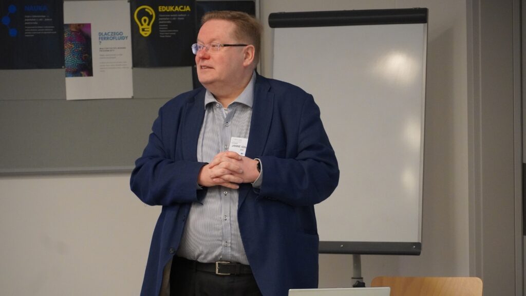Speaker presenting during a conference session, standing in front of a flip chart and informational posters, addressing an audience in a meeting room.