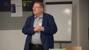 Speaker presenting during a conference session, standing in front of a flip chart and informational posters, addressing an audience in a meeting room.