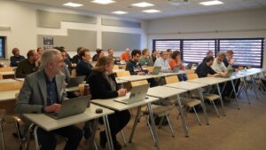 Conference attendees seated in a classroom-style setting, working on laptops and listening during a session in a meeting room.
