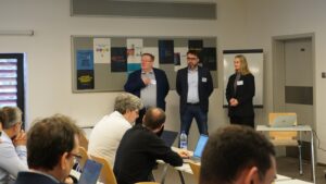 Three speakers standing at the front of a meeting room addressing attendees seated with laptops, during a conference session with posters displayed on the wall behind them.