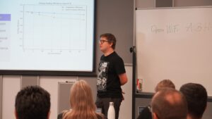 Speaker presenting technical content during a conference session, standing beside a projected chart on scaling efficiency while addressing an audience.