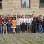 Large group of conference participants standing outdoors in front of a stone building, posing together for a group photo in daylight.
