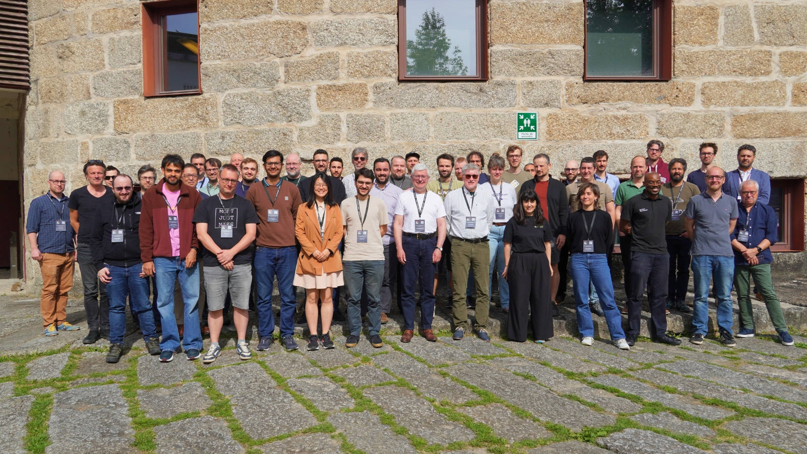 Large group of conference participants standing outdoors in front of a stone building, posing together for a group photo in daylight.