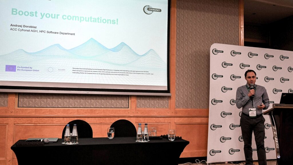 Speaker presenting “Boost your computations!” on stage at a Cyfronet event, standing beside a projected slide with EuroHPC and EU co-funding logos visible.