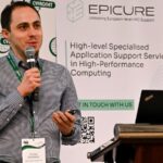 Speaker holding a microphone and presenting at an EPICURE stand, with a banner about specialised HPC application support visible in the background.