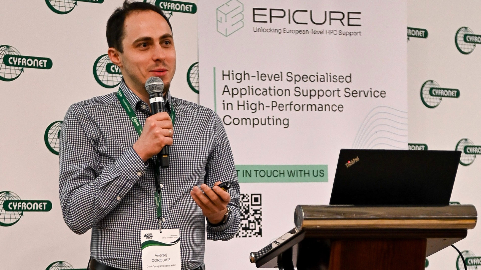 Speaker holding a microphone and presenting at an EPICURE stand, with a banner about specialised HPC application support visible in the background.