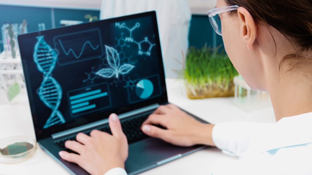 Researcher working on a laptop displaying scientific data visualisations, including DNA structure, molecular diagrams, and charts, in a laboratory setting.