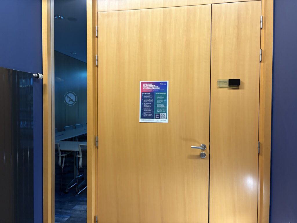 Closed wooden meeting room door with a poster attached, next to a glass panel showing a conference room with chairs inside.