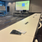 Empty training room with U-shaped tables, a large presentation screen displaying a welcome slide, and EPICURE materials placed on the table in the foreground.