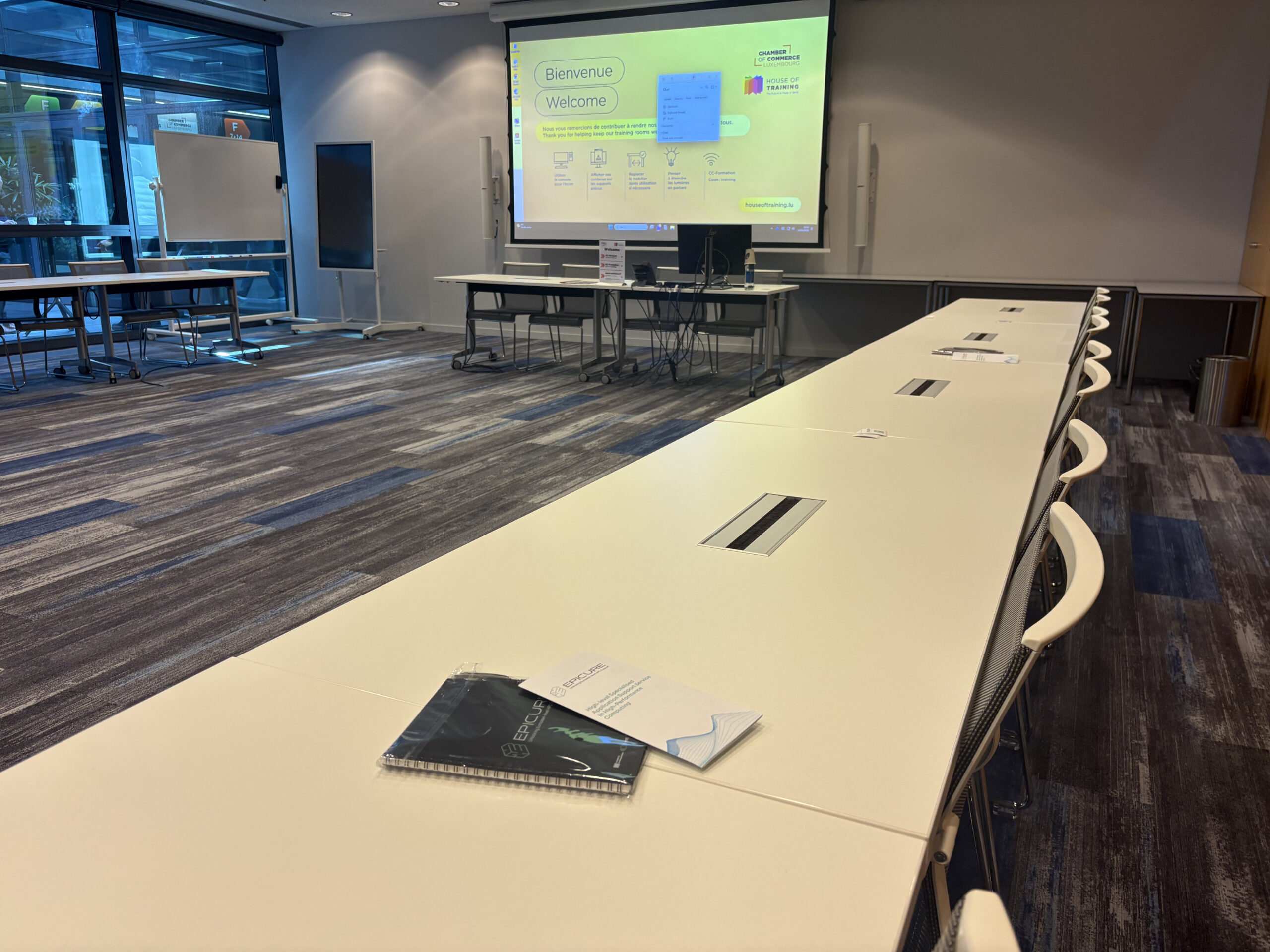 Empty training room with U-shaped tables, a large presentation screen displaying a welcome slide, and EPICURE materials placed on the table in the foreground.