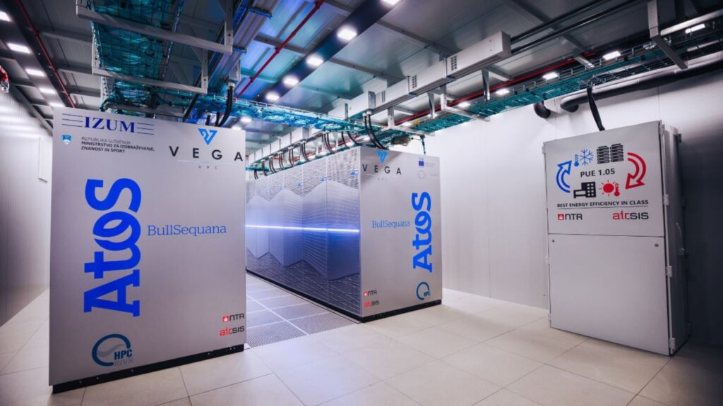 Alt text (EN-UK): High-performance computing system inside a data centre, featuring Atos BullSequana cabinets from the VEGA supercomputer, with visible cooling infrastructure and energy efficiency signage.