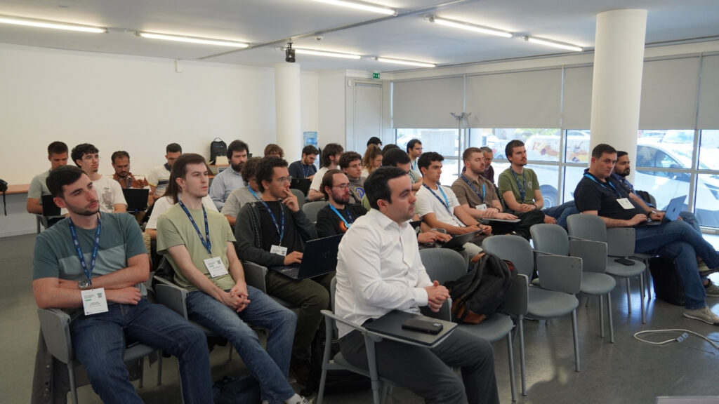 Audience seated in a workshop room, listening attentively to a technical presentation, with several participants using laptops.