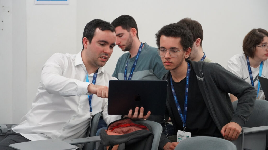 Participant guiding a colleague through content on a laptop during a hands-on HPC workshop session, with other attendees working in the background.