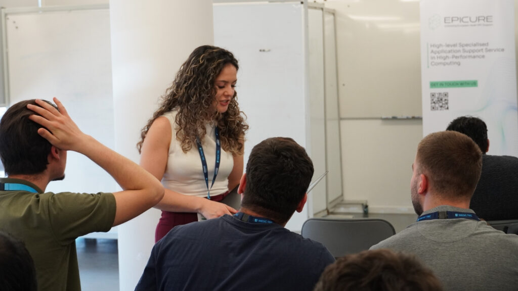 Facilitator assisting participants during a hands-on HPC workshop, discussing work on a laptop while attendees listen and engage.