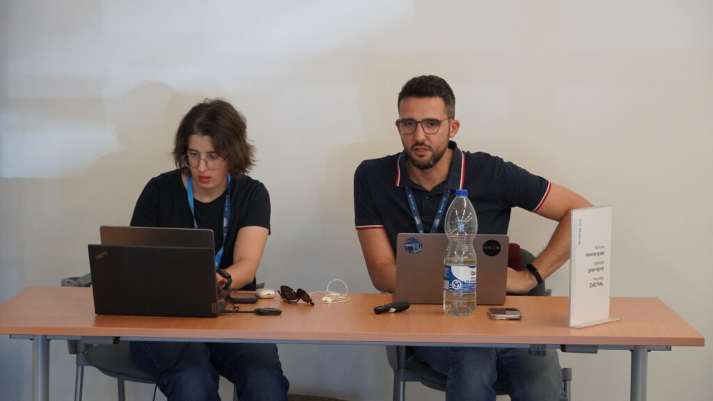 Two EPICURE Hackathon organisers seated at a registration and support desk with laptops and event materials, assisting participants during the HPC optimisation event at INESC TEC.