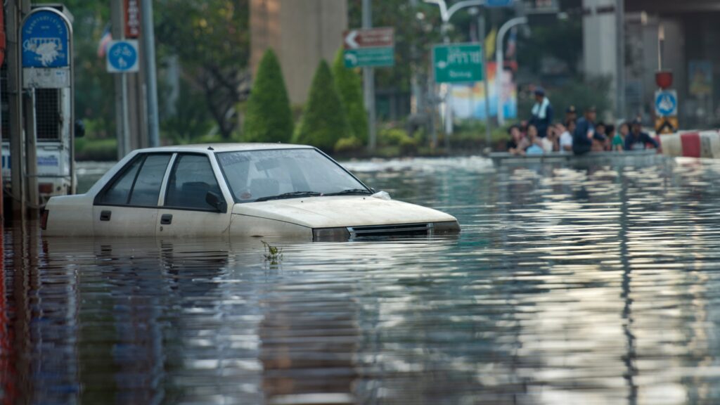A partially submerged car sits in a flooded urban street, surrounded by high water levels that have disrupted traffic and infrastructure. The image illustrates the severe impact of flooding events on cities, transportation and public safety.